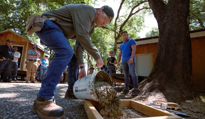 A person in work boots pours a bucket of concrete into a wooden form on the ground.