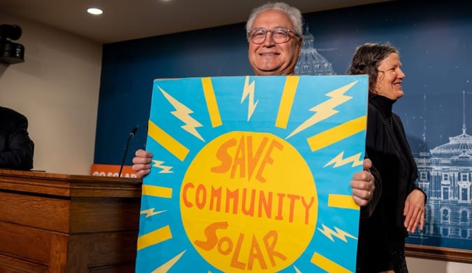 Man near podium indoors holds hand-painted blue sign withe drawing of a sun that says "Save Community Solar"