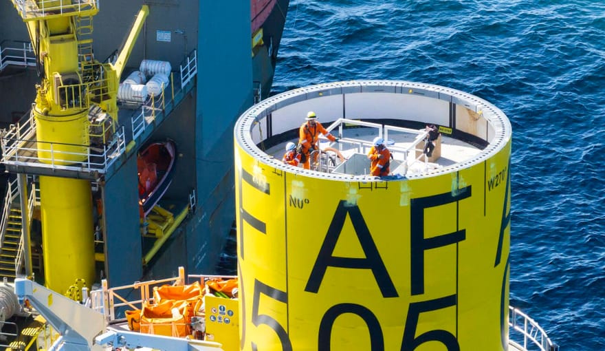 Workers on a large yellow cylinder in front of ocean water