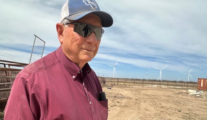 A person wearing a button-up shirt, sunglasses, and a hat stands outside with wind turbines behind him.