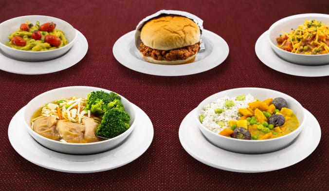 Five plant-based meals on white plates against a plain red background.