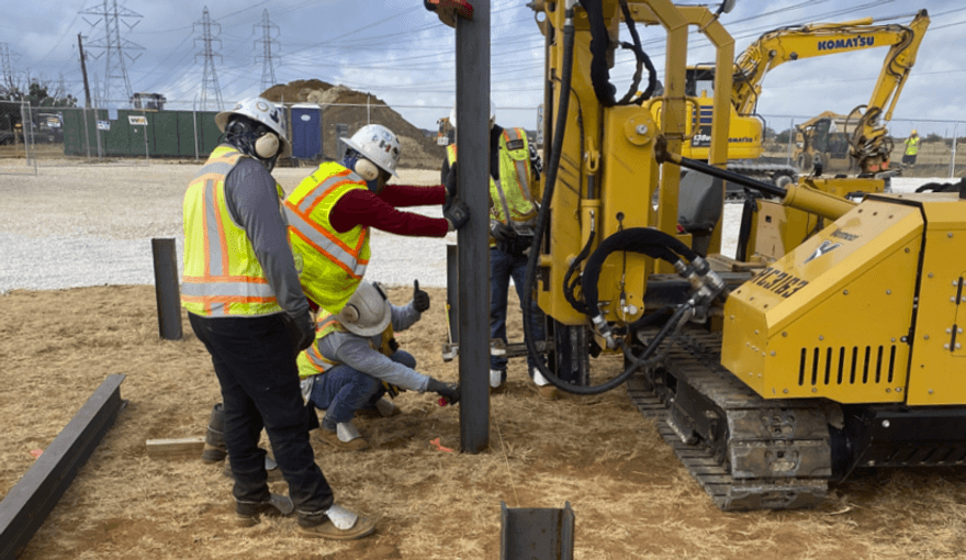 Several workers in neon yellow vests and hard hats surround a piece of machinery drilling into the ground