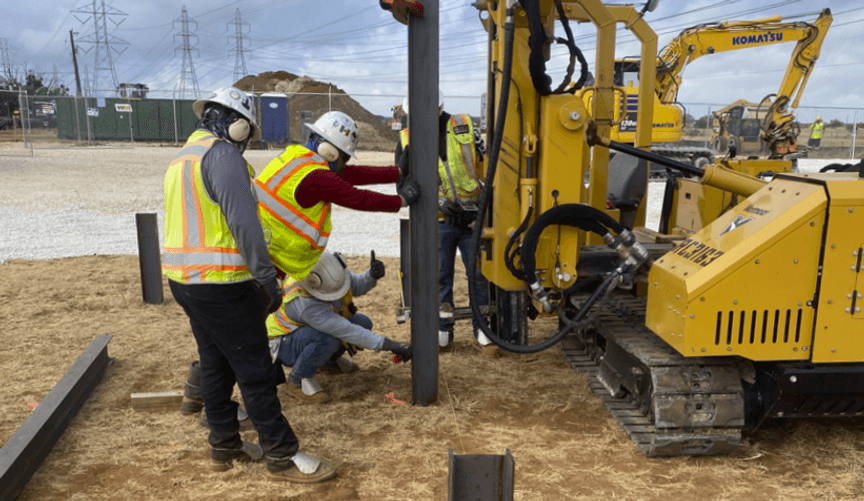 Several workers in neon yellow vests and hard hats surround a piece of machinery drilling into the ground