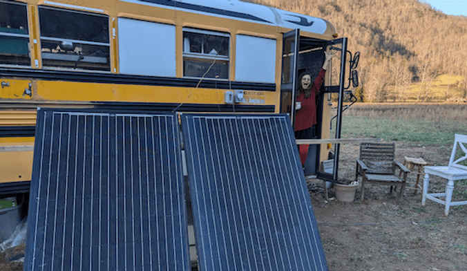 A person stands in the door of a school bus. Two solar panels lean against the outside of the bus.